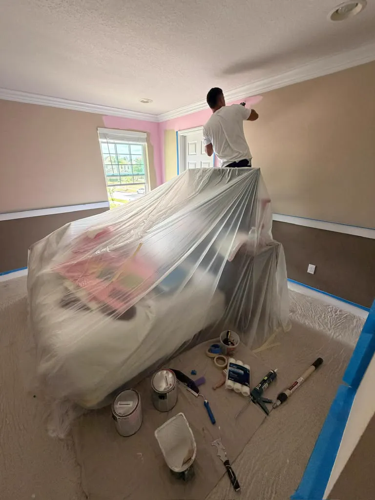 A bedroom with the furniture wrapped in plastic while the walls are painted